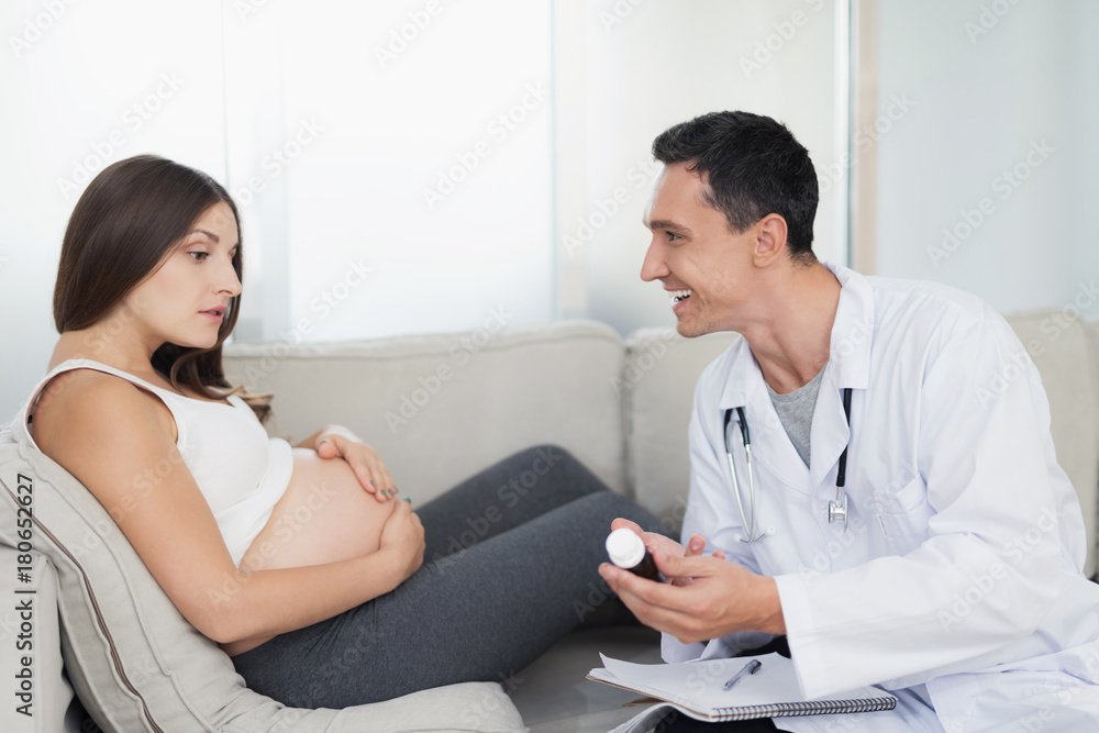 A pregnant woman lies on a light sofa at home. A doctor came to her, he offers her some kind of medical product A pregnant woman lies on a light sofa at home. A doctor came to her, he offers her some kind of medical product