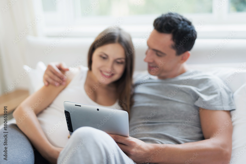 A pregnant woman and a man are lying on the bed. They are looking at something on their tablet A pregnant woman and a man are lying on the bed. They are looking at something on their tablet