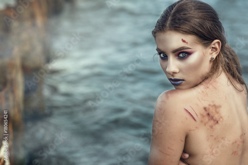 Portrait of young beautiful scared woman standing in the misty sea water and looking over her shoulder. Her back covered with scratches and bruises. Gender based violence concept. Outdoor shot