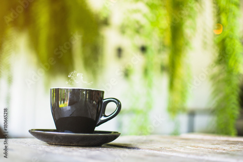 Black cup with steaming hot coffee on the wooden table with blurred green garden background