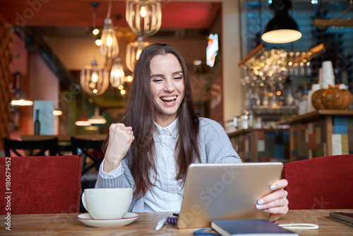 successful young beautiful dark-haired woman with long hair sitting in a cafe with a tablet with a happy face clutching a fist in the pose of a winner.