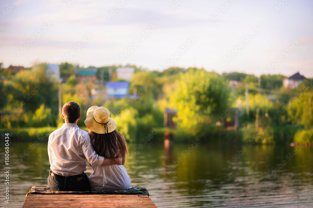 Beautiful bright fairy walk of a loving couple in the summer at sunset. A guy in a white shirt and jeans with a girl in a sarafan embraces, dancing, sitting on the pier. Pond in the background. Place Beautiful bright fairy walk of a loving couple in the summer at sunset. A guy in a white shirt and jeans with a girl in a sarafan embraces, dancing, sitting on the pier. Pond in the background. Place