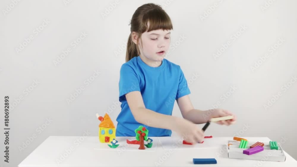 Smiling girl in blue molds house from plasticine on white table in white room
Smiling girl in blue molds house from plasticine on white table in white room