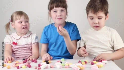 Three children eat candies and sit at table in white studio
