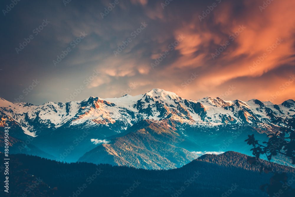 Beautiful colorful sunset over the snowy mountain range and pine tree forest. Nature landscape. Dramatic overcast sky with orange clouds. Main Caucasian ridge, Svaneti, Georgia. Retro toning filter