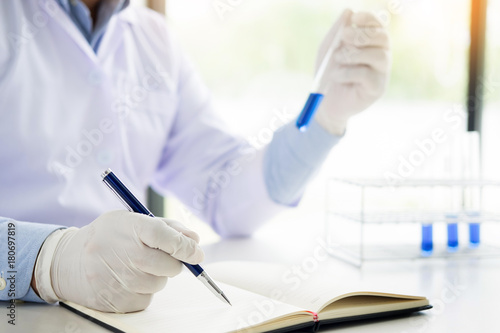 Scientist in white coat holding and examining test tube with reagent making notes of his research in laboratory