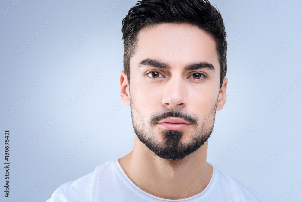 Nice portrait. Young handsome man with a black beard looking calm and concentrated while standing against the blue background Nice portrait. Young handsome man with a black beard looking calm and concentrated while standing against the blue background