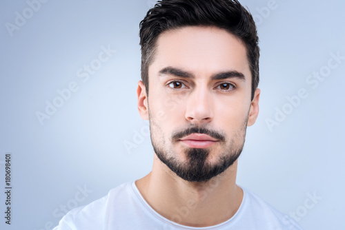 Nice portrait. Young handsome man with a black beard looking calm and concentrated while standing against the blue background