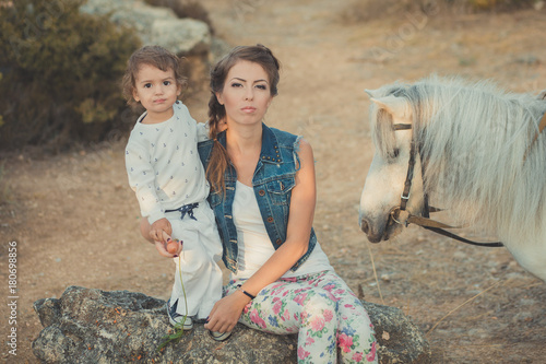 Romantic scene beautiful lady young mother with her cute baby daughter enjoy time together in city village park walking feeding white horse pony photo