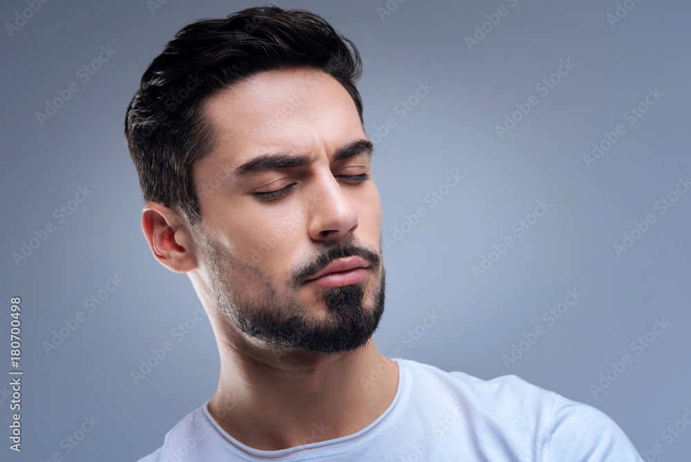 Feeling calm. Relaxed unemotional young man sitting with his eyes closed and meditating while being absolutely alone at home Feeling calm. Relaxed unemotional young man sitting with his eyes closed and meditating while being absolutely alone at home