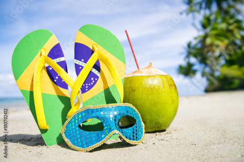 Brazil flag flip flops, colorful sequined carnival mask and fresh green coconut drink on a tropical palm fringed beach.