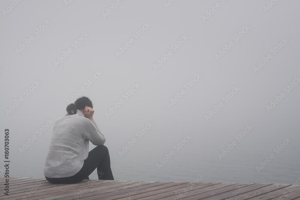 Junge Frau sitzt einsam am Rand eines Holzweges einer Brücke gebückt und traurig in Gedanken versunken im Nebel Junge Frau sitzt einsam am Rand eines Holzweges einer Brücke gebückt und traurig in Gedanken versunken im Nebel