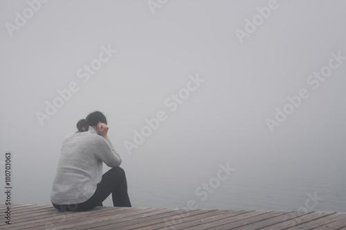 Junge Frau sitzt einsam am Rand eines Holzweges einer Brücke  gebückt und traurig in Gedanken versunken im Nebel photo