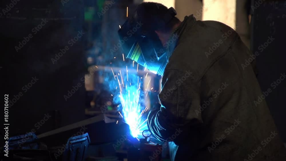 A laborious man in a uniform and a black protection mask uses a welding machine, making sparkling blue light. He checks his work. The whole workshop looks dark and slightly lit. A laborious man in a uniform and a black protection mask uses a welding machine, making sparkling blue light. He checks his work. The whole workshop looks dark and slightly lit.
