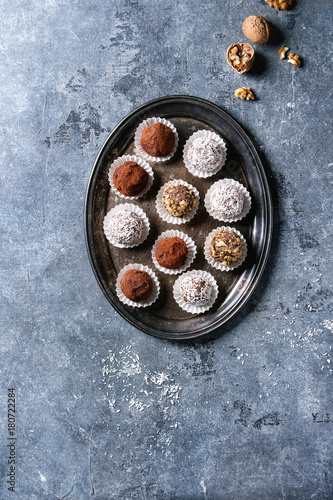 Variety of homemade dark chocolate truffles with cocoa powder, coconut, walnuts on vintage tray over blue texture background. Top view, copy space.