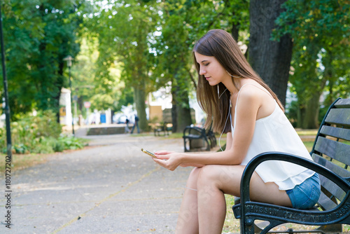 Young smiling woman with long hair in summer park sits on a bench and use smartphone