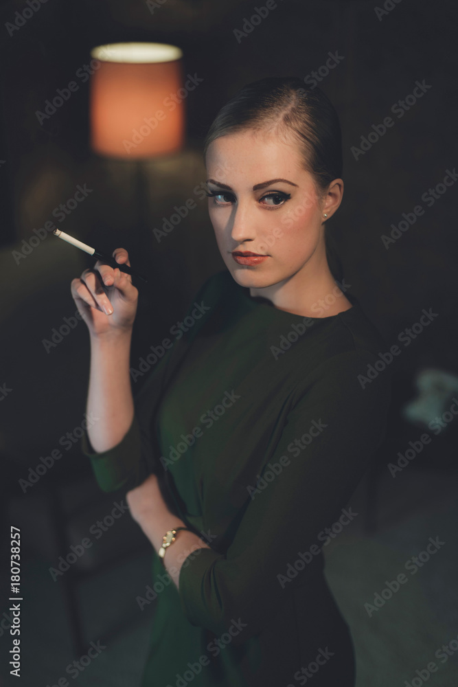 Obraz premium Retro woman in green dress standing with cigarette in hotel room at night.