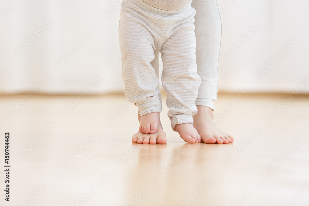 Fototapeta premium Cute baby boy learning to walk and make his first steps. mom is holding his hand. child's feet close up, copy space