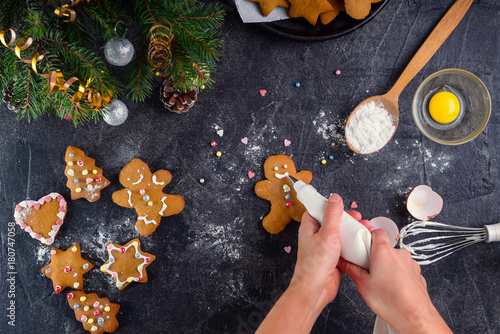Woman hands holding a culinary bag with meringue cream inprocess of decorating gingerbread man cookies. Black stone background with ingrediens and decoration details. Christmas mood concept.