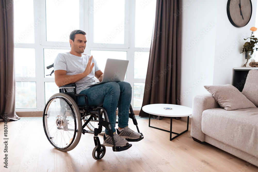 The disabled person sits in a wheelchair in front of a large panoramic window. He has a laptop on his lap, for which he works. He is in his large bright living room. The disabled person sits in a wheelchair in front of a large panoramic window. He has a laptop on his lap, for which he works. He is in his large bright living room.