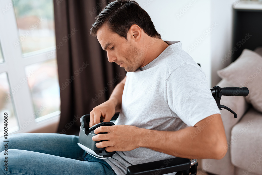 A disabled person in a wheelchair is going to put on a virtual reality glasses. He sits in his large bright living room. He wears a light T-shirt and jeans.