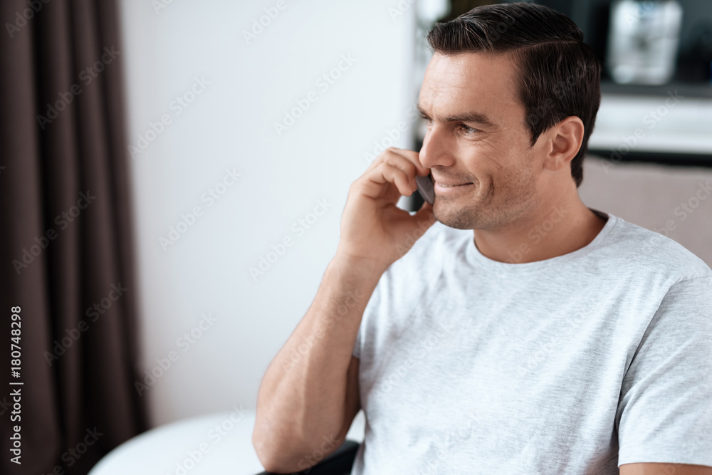 The disabled person sits in a wheelchair in front of a large panoramic window. He is talking to someone on his smartphone. He is in his large bright living room. He smiles. The disabled person sits in a wheelchair in front of a large panoramic window. He is talking to someone on his smartphone. He is in his large bright living room. He smiles.