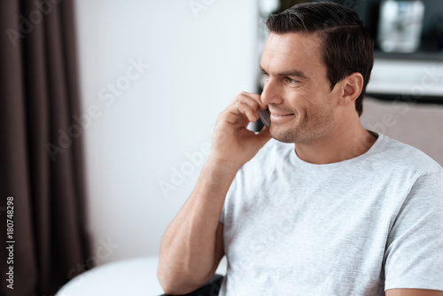 The disabled person sits in a wheelchair in front of a large panoramic window. He is talking to someone on his smartphone. He is in his large bright living room. He smiles.