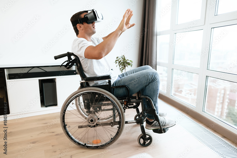 Obraz premium The disabled person is sitting in a wheelchair in virtual reality glasses. He spread his hands to the sides. He sits in his large bright living room and enjoys virtual reality.