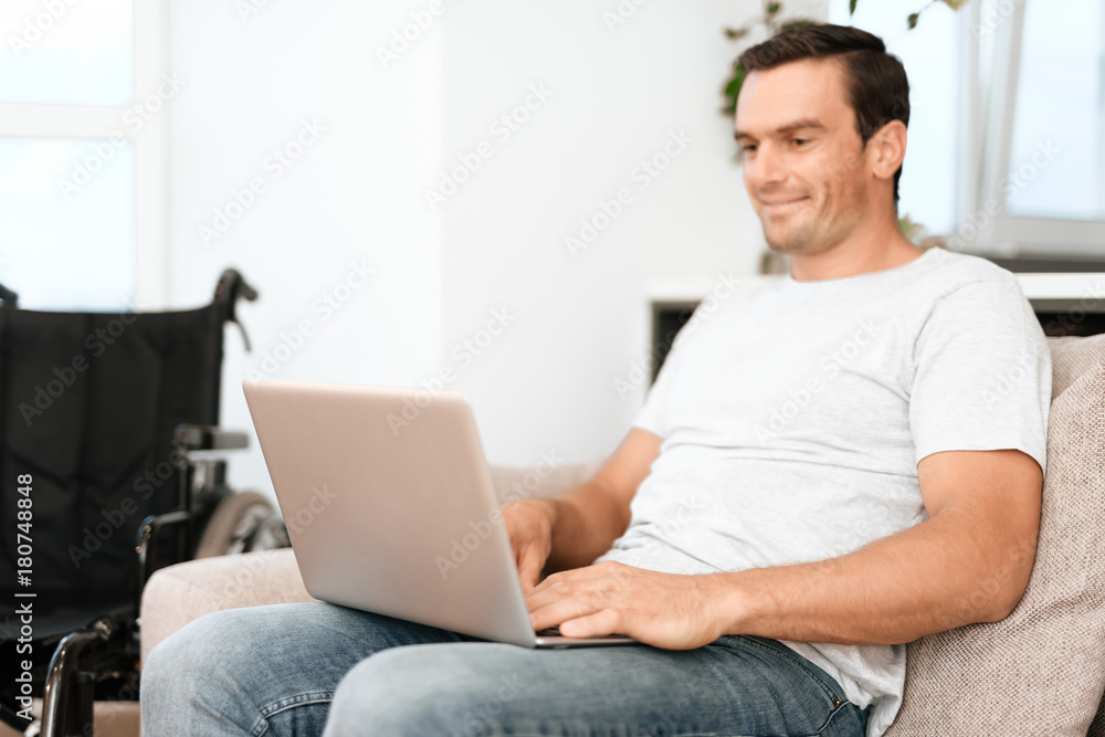 The disabled person is sitting on the couch. He works for his laptop. next is his wheelchair. He sits on a large bright couch in his modern apartment. The disabled person is sitting on the couch. He works for his laptop. next is his wheelchair. He sits on a large bright couch in his modern apartment.