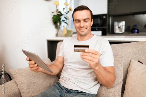 The disabled person is sitting on the couch. He has a tablet and a credit card in his hands. He wants to buy something online. The man is smiling. Next to him is his wheelchair.