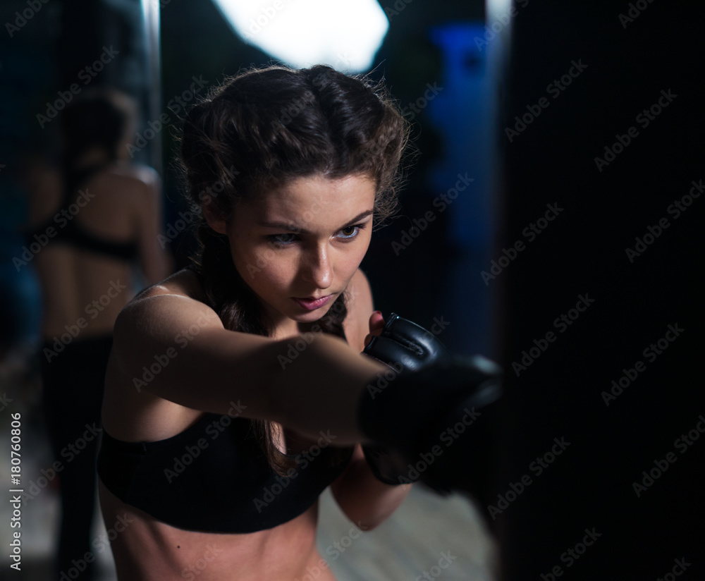 Beautiful young fighter boxer fit girl wearing boxing gloves in training, focused on heavy punching bag in gym. She is in good shape. Woman power