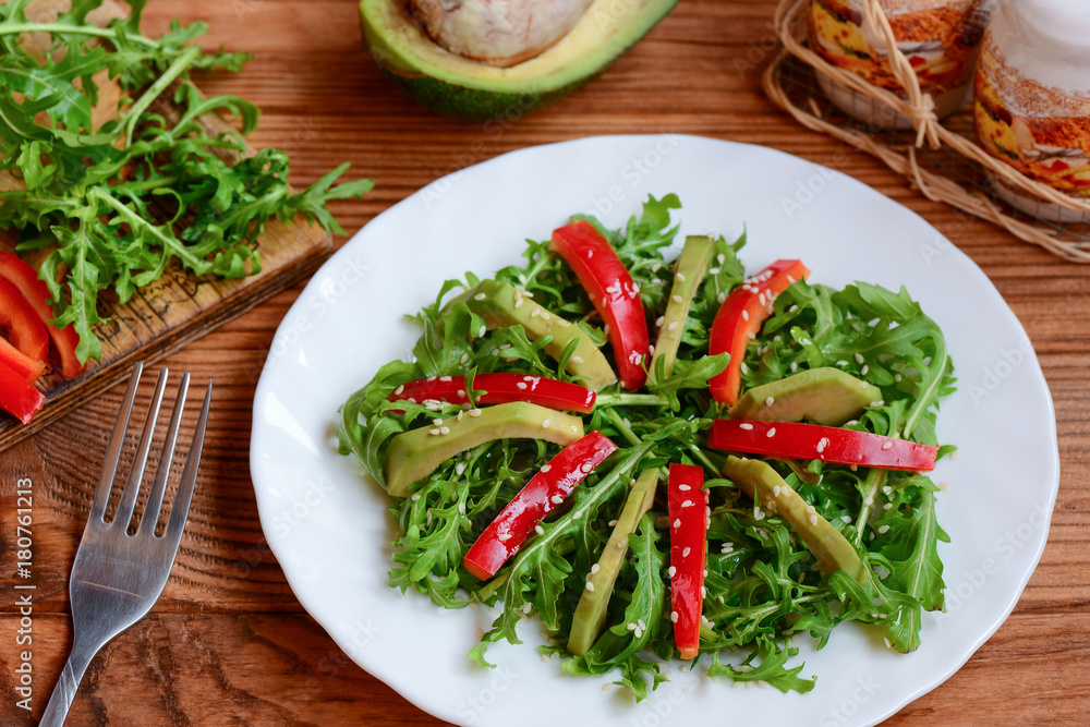 Fresh avocado, arugula and pepper salad. Easy arugula and avocado salad with fresh pepper and sesame seeds on a white plate and wooden table. Raw food diet. Closeup