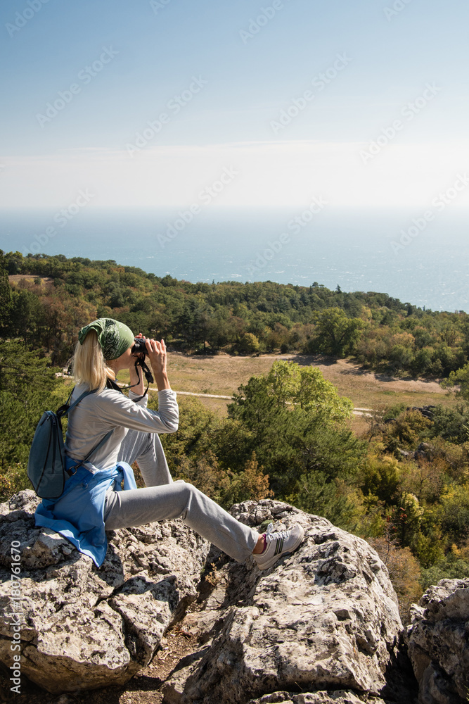 A blond Woman tourist with a backpack on a cliff looking through binoculars on the sea. Travel concept.