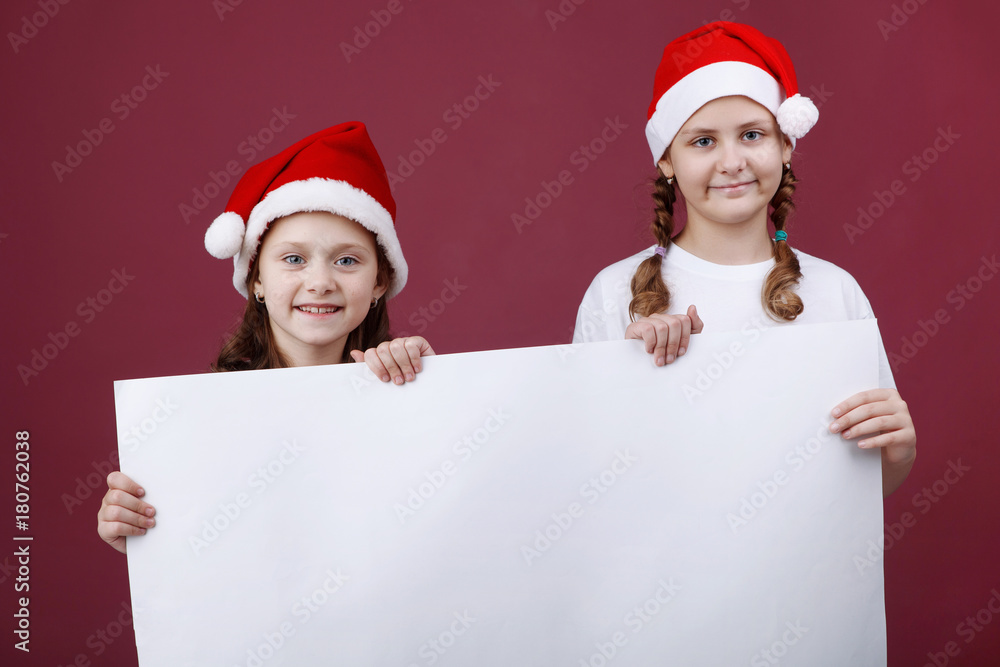 Two smiling little girl in hat of Santa Claus with white blank banner isolated on red. Two smiling little girl in hat of Santa Claus with white blank banner isolated on red.