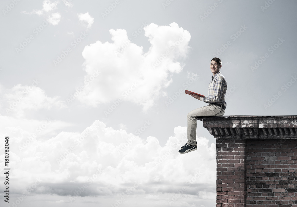 Man on roof edge reading book and cloudscape at background Man on roof edge reading book and cloudscape at background