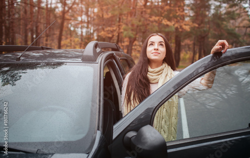Long-haired brunette on the auto background. A female model is wearing a sweater and a scarf. Autumn concept. Autumn forest journey by car