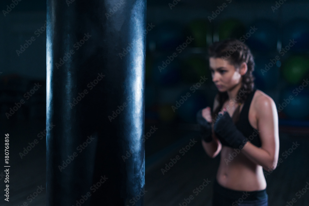 Young fighter boxer fit girl wearing hand bandage in training with heavy punching bag in gym in bokeh. Focus on punching bad