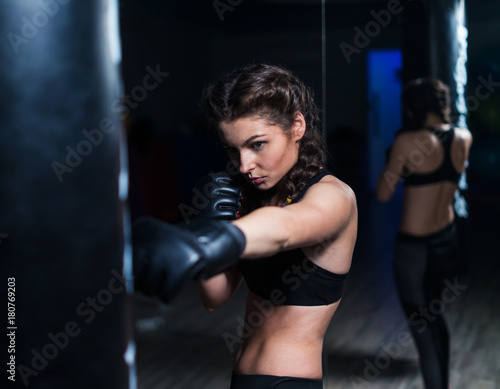 Cute fighter boxer fit girl wearing boxing gloves in training with heavy punching bag in gym. She is in good shape. Woman power