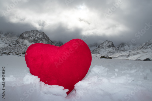 Red plush heart in snow on a background of snow-capped mountains.