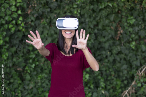 Young beautiful woman enjoying virtual reality glasses headset or 3d spectacles over green ivy background. Outdoors. Technology, innovation, cyberspace and gaming. © Eva