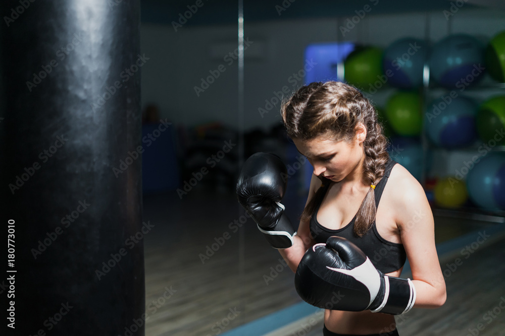Upset young fighter boxer girl wearing boxing gloves in gym beating punching bag. She is sad