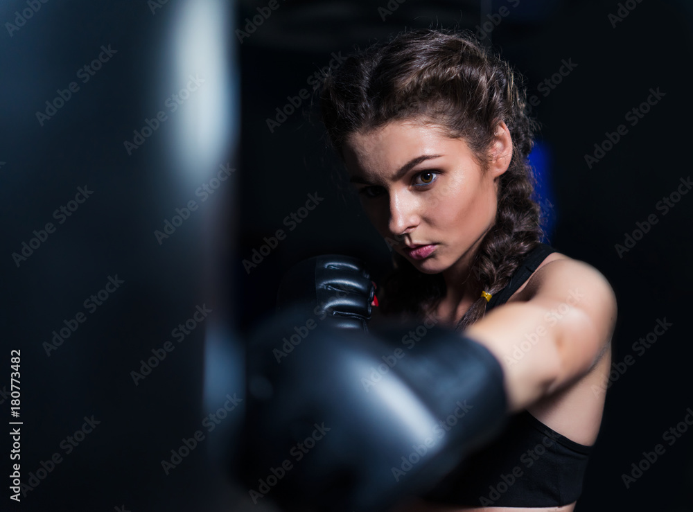 Beautiful fighter boxer fit girl wearing boxing gloves in training with heavy punching bag in gym. Low key image. Woman power