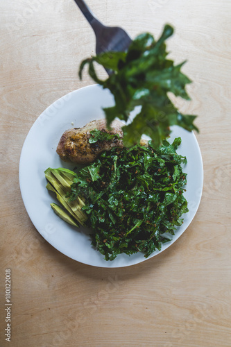 Organic baby kale green salad tossed with gluten free bread crumbs and plated with an organic roasted chicken leg and half of an organic avocado on a white plate against a wooden background. photo