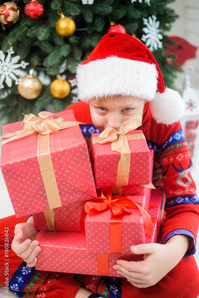 Portrait of cute funny little kid dressed in red night-suit and hat of santa. White happy kid holding stack of holiday presents in hand and smiles happily. Vertical color photo.