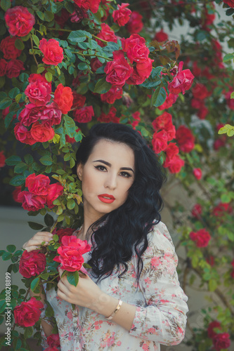 Amazing mystic lady woman with black dark curly hairs pink cheeks pout deep red lips wearing designer couturier shirt and blue short jeans shorts with brown belt posing sit for bush of flowers roses