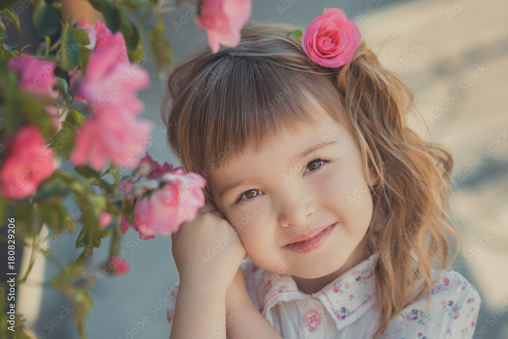 Cute baby girl with blond curly hairs and happy bright child eyes posing in central park close to huge bush of flowers red white pink roses wearing summer stylish clothes Cute baby girl with blond curly hairs and happy bright child eyes posing in central park close to huge bush of flowers red white pink roses wearing summer stylish clothes
