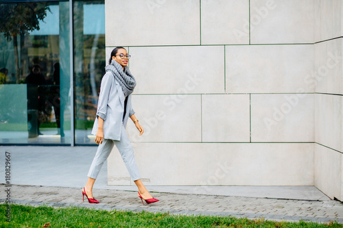 Side view full body of african business woman in gray suit going along beige wall of business center. Lifestyle, leisure and people concept. photo