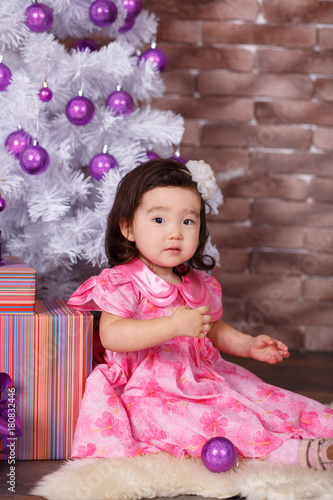Asian pin up little cute girl wearing pink casual dress posing close to new year christmas white tree with violet purple balls toys and colourful presents