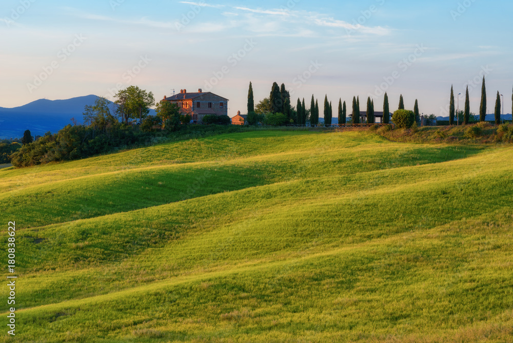 Fototapeta premium Magnificent spring landscape at sunrise.Beautiful view of typical tuscan farm house, green wave hills, cypresses trees, hay bales, olive trees, beautiful golden fields and meadows.Italy, Europe