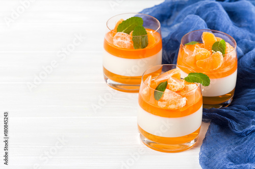 Creamy panna cotta with orange jelly in beautiful glasses, fresh ripe mandarin, blue textile on white wooden background. Delicious Italian dessert. Closeup photography.Selective focus.